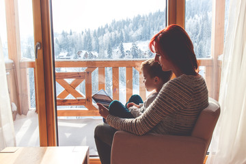 Mother and her son sitting in a comfortable chair with tablet next to the window with look at snow...