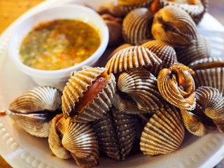 Thai local raw fresh red bloody semi short time steamed blanched open cockle clam pile on white foam plate, with blurred spicy seafood chili dipping sauce background, under night yellow lighting
