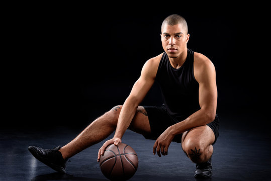 handsome african american basketball player sitting with ball on black