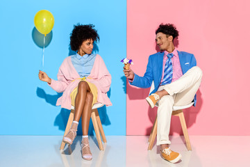 african american man presenting flowers in ice cream cone to girlfriend while sitting on chairs against pink and blue wall backdrop