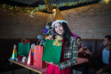 Obraz na płótnie Canvas beautiful woman wear red dress and santa claus hat showing green gift bag on hand in restaurant. concept of Christmas party and New year party.