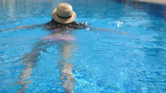 Back of young unrecognizable girl in hat swimming in pool. Woman relaxing in basin. Holiday concept. Rear view Close up Slow motion