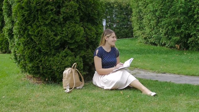 Beautiful young woman sitting in park on grass and writing a diary. Slow motion footage