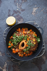 Osso buco with gremolata in a cast-iron pan on a metal cooling rack. Top view on a brown stone background with copyspace