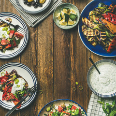 Flat-lay of healthy dinner table setting. Fresh salad, grilled vegetables with yogurt and dill sauce, pickled olives, lemon water over wooden background, copy space, square crop. Clean eating