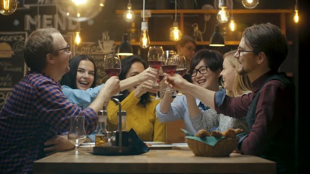 Diverse Group of Friends Celebrate with a Toast and Clink Raised Wine Glasses in Celebration. Beautiful Young People Have Fun in the Stylish Bar/ Restaurant. 