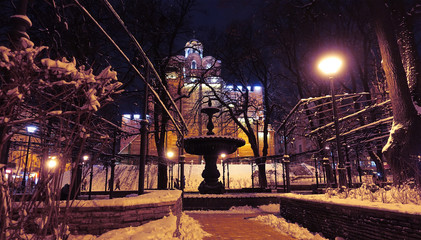 An ancient fountain in the park near the Golden Gate in Kiev. Winter snowy evening. Lights illuminate the scene