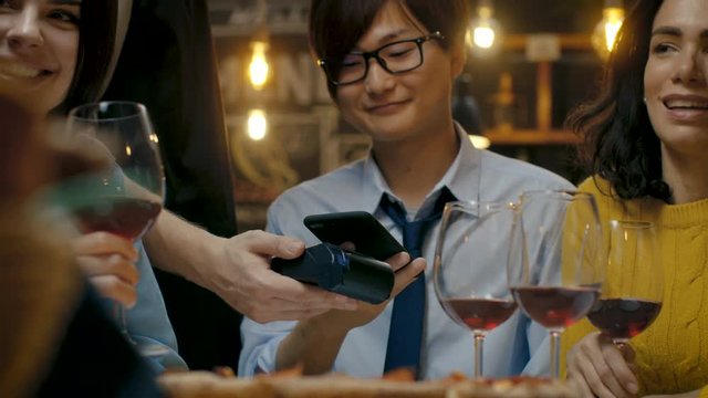 In the Bar Waiter Holds Credit Card Payment Machine and Man Pays for His Order with Contactless Mobile Phone Payment System. He's Surrounded by Friends and Has Great Time. 