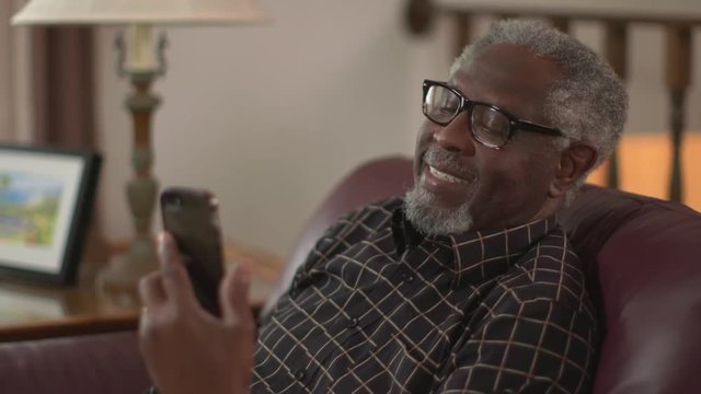 A happy elderly African American grandpa smiling and waving, chatting on a smartphone or iPhone. Authentic family feel. Slow motion (48fps) Prores file.