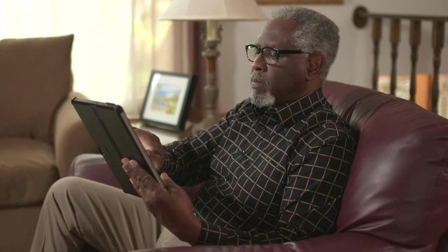 Elderly African American retiree online using a tablet computer, reading. Authentic at home lifestyle shot. Social distancing. Prores file.