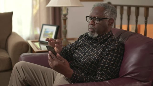 Elderly African American Retiree Using A Smartphone Or IPhone To Browse Online And Read. Authentic At Home Lifestyle Shot. Prores File.