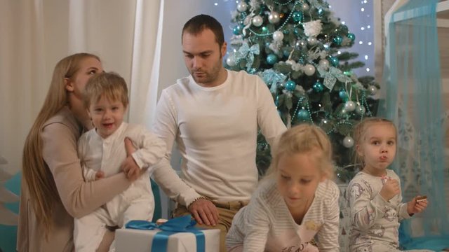 A happy family sitting by the New Year tree, parents are playing with children, smiling and laughing