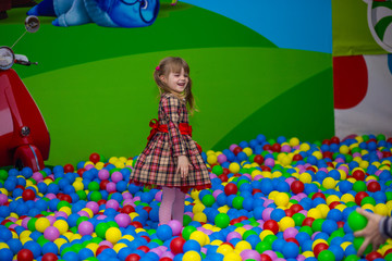 Fototapeta na wymiar Happy little kid girl in red dress play in pool with colorful plastic balls. Funny child having fun indoors.