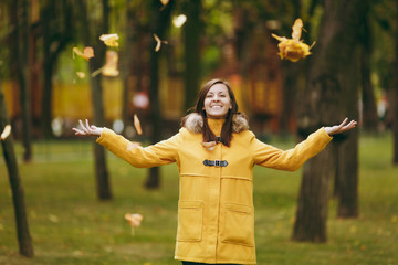 Beautiful happy caucasian young smiling brown-hair woman in yellow coat, jeans, boots in green forest. Fashion female model throwing up fall leaves standing and walking in early autumn park outdoors.