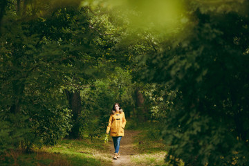 Fototapeta premium Beautiful happy caucasian young smiling brown-hair woman in yellow coat, jeans, boots in green forest. Fashion female model with fall golden leaves standing and walking in early autumn park outdoors.