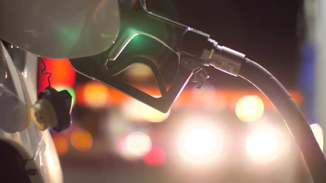 Fuel nozzle inserted in car's gas tank as it's being refueled at gas station pump at night. Closeup, shallow DOF. 4K UHD