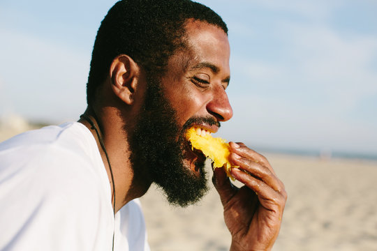 Man with beard and dark skin eating mango on sea shore