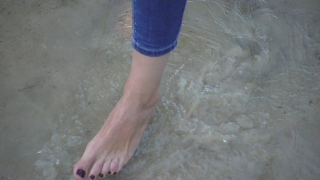 Female bare foot standing and moving sea water on background sandy bottom slow motion. Close up woman leg muddy clear water and sand on sea coast
