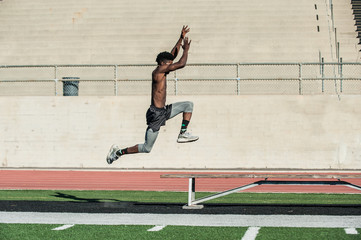 Jamaican track athlete performing explosive long jump over bench at stadium. 