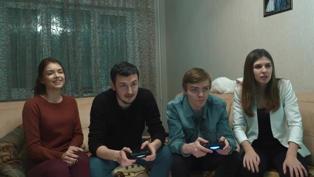 Two guys and two girls play on the game console. They are sitting on the couch and holding the joysticks. One of the guys is disabled in a wheelchair
