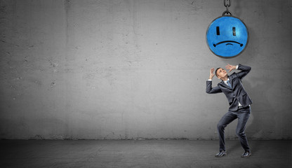 Obraz premium A scared businessman stands on concrete background under a wrecking ball with a painted blue sad face.