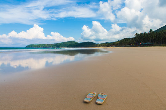 pair of flipflops in empy sand beach with cloudy sky and tropical trees on background