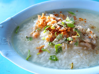Thai local traditional white Jasmine rice fully boiled duck meat porridge with spring onion, dry cabbage leaves, and pepper powder in long oval blue bowl, close up top view, on old table