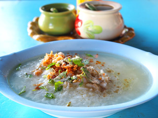 Thai local traditional white Jasmine rice fully boiled duck meat porridge with spring onion, dry cabbage leaves, and pepper powder in long oval blue bowl, ceramic cups of seasoning sauce on a tray
