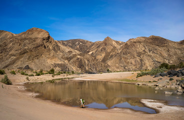 A lone hiker walking along a large pool of water on the Fish River while hiking through the arid...