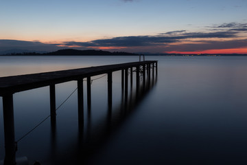 Long exposure view of a pier on a lake at dusk, with distant colorful clouds and perfectly still water
