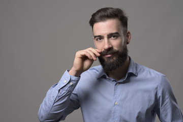 Moody portrait of young stylish bearded man twirling mustache and looking at camera over gray studio background with copyspace. 