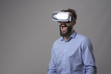 Portrait of amazed excited young bearded man watching virtual reality glasses with open mouth expression over gray background with copyspace. 