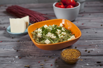 pasta green with spinach and mozzarella cheese and greens in an orange bowl, French mustard, colored raw spaghetti, saucer with cheese, dill and bowl with cherry tomatoes on a light wooden table
