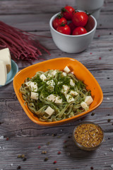 pasta green with spinach and mozzarella cheese and greens in an orange bowl, French mustard, colored raw spaghetti, saucer with cheese, dill and bowl with cherry tomatoes on a light wooden table