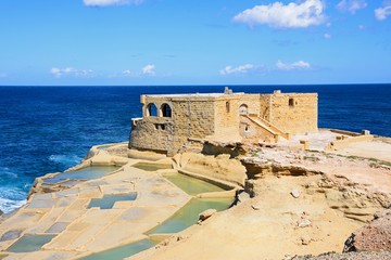 Salt pans along the waterfront with the Il-Qolla I-Badja battery to the rear, Marsalforn, Gozo, Malta.