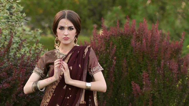 Beautiful hindu girl in traditional costume sari of dark red color on the background of green grass and looking at the camera. Girl with pictures of mehendi on the hands and kundan jewelry.