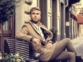 Handsome man wearing elegant coat sitting on bench at street and posing relaxed. 
