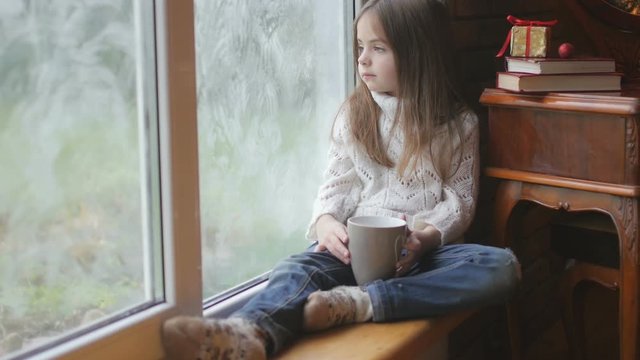 beautiful little girl with a mug of hot tea sits by the window, looks at the street and what she dreams about