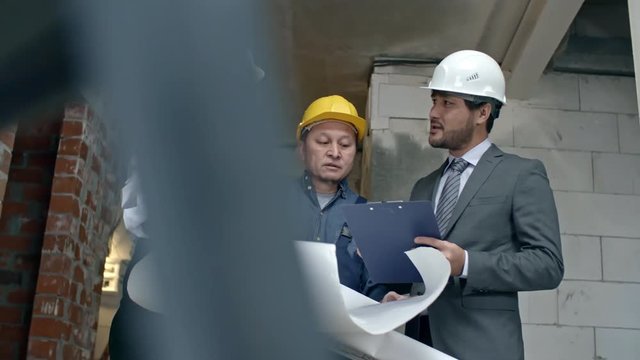 Low angle PAN of female construction contractor in hard hat showing blueprints and discussing work with Asian businessman with clipboard and male builder in blue overalls while inspecting building