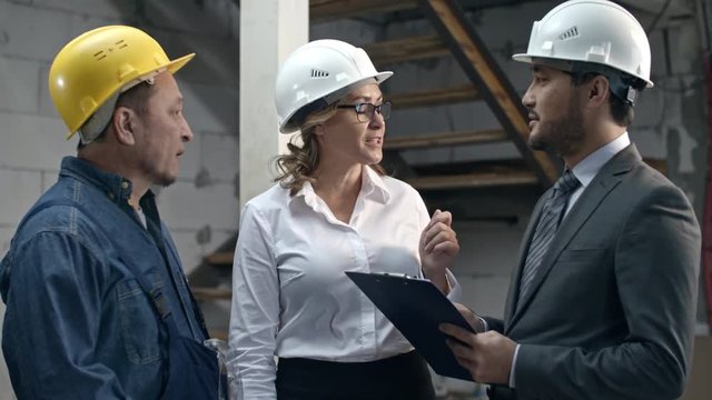Slowmo of businesswoman in hard hat standing inside unfinished building and discussing completion date of construction with Asian businessman with clipboard and foreman holding paintbrush
