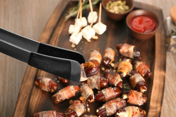 Holding tasty bacon wrapped date with tongs over wooden tray