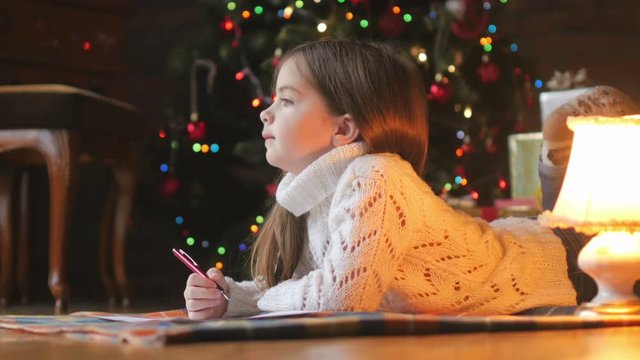beautiful little girl in a white knitted sweater lies on the floor and writes a letter to Santa Claus, in the background a festive Christmas tree and a lot of presents