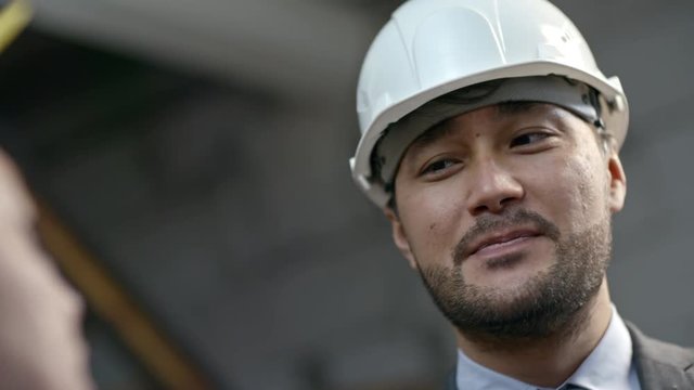 Close up with PAN of cheerful Asian supervisor in hard hat talking to unrecognizable male factory worker