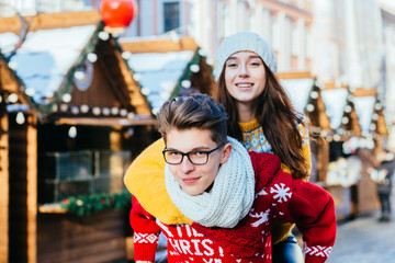 Happy winter travel couple. Guy giving girlfriend piggyback ride on winter vacation in Christmas fair market at central square of the european city. Active lifestyle, vacation and relationship concept