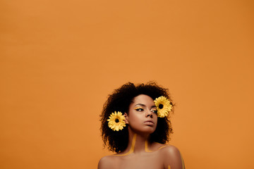 Young sensual african american woman with artistic make-up and gerbera in hair isolated on orange...