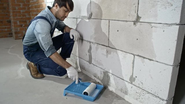 Tilt up of Asian construction painter in blue overalls putting priming coat on brick wall with paint roller