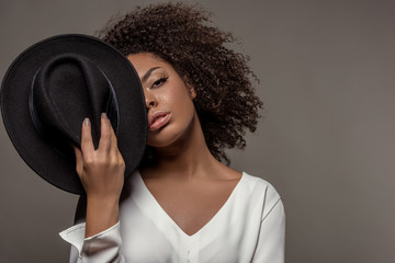 Attractive young african american woman in white shirt holding black hat over half of her face isolated on grey background - obrazy, fototapety, plakaty