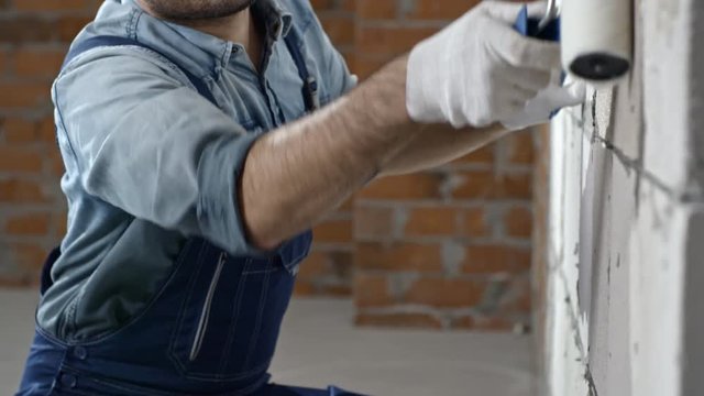 Tilt down of cheerful male Asian construction painter putting priming coat on brick wall with paint roller