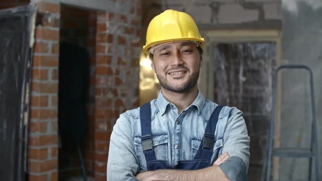PAN of cheerful young Asian builder in hard hat standing with crossed arms in unfinished building and smiling for camera