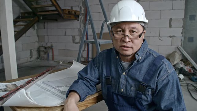 Medium shot of mature Asian engineer wearing glasses and hard hat sitting in unfinished building and explaining something while looking at camera